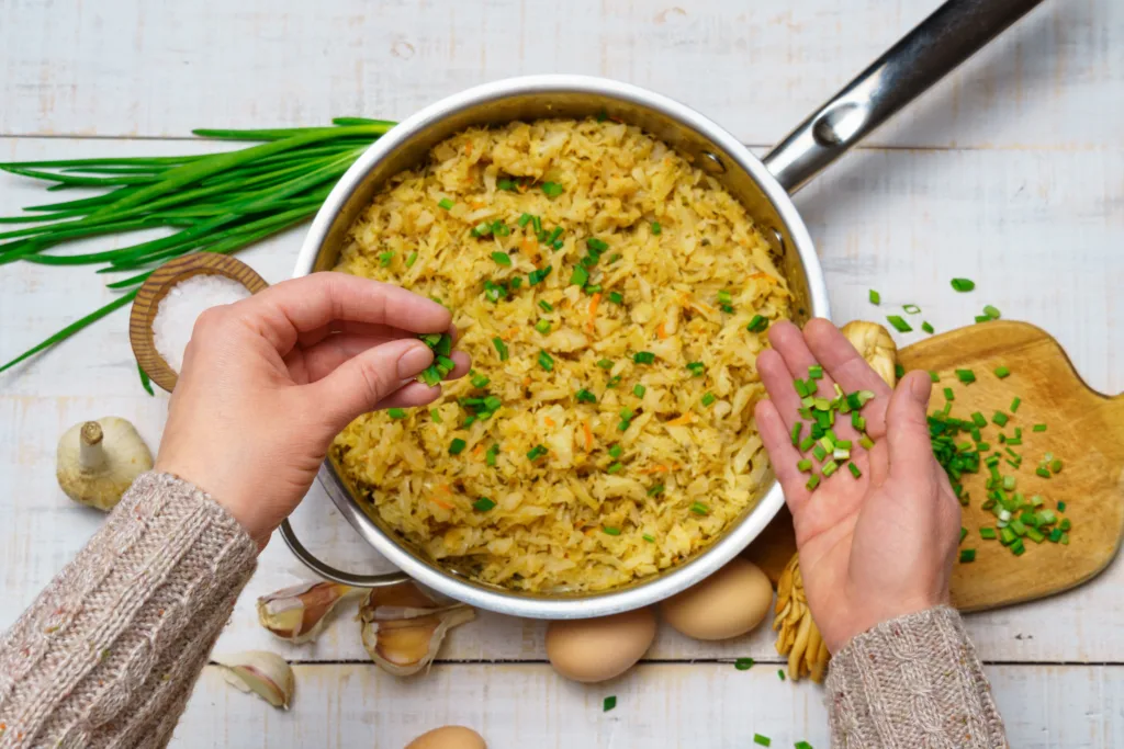 a woman's hand takes fried chopped cabbage in a frying pan, garlic and green onions, cheese, farm eggs and salt, raw and cooked food in a rustic style on a background of white wooden boards