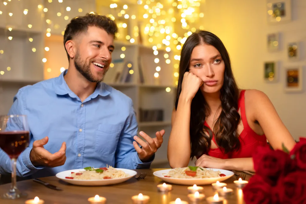 At candlelit dinner, cheerful man speaks animatedly while woman in red dress appears unimpressed, resting her cheek on her hand, sitting at dinner table