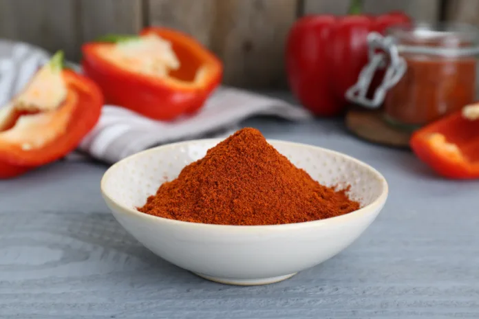 Bowl with aromatic paprika powder and fresh bell peppers on grey wooden table