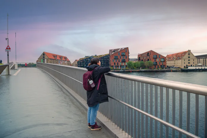 brunette girl in warm clothes looks leaning on handrails Inner Harbour pedestrian and bicyclist bridge Inderhavnsbroen against the neighbourhood Christianshavn