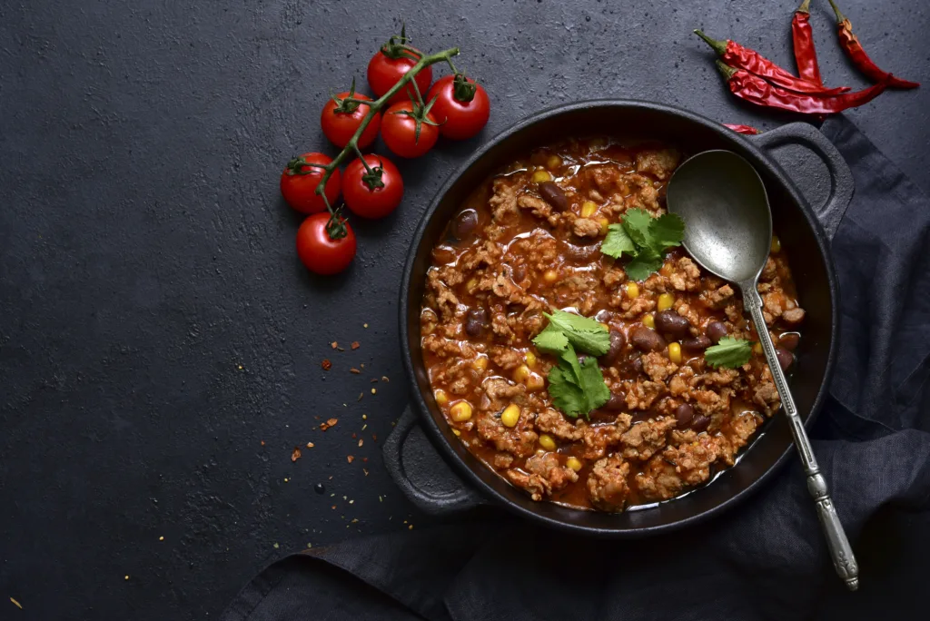 Chili con carne - traditional mexican minced meat and vegetables stew in tomato sauce in a cast iron pan on a black slate, stone or concrete background. Top view with copy space.