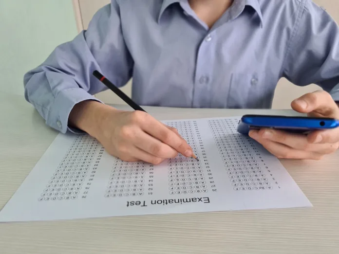 Closeup of teenager sitting at desk holding phone in her hand and taking exam test. Focused guy preparing for test or college exam