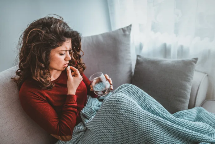 Concept of suffering from terrible pain. Beautiful, sad, upset, unhappy, troubled, weak woman with blonde hair, wearing casual clothes is sitting on a sofa and taking a pill