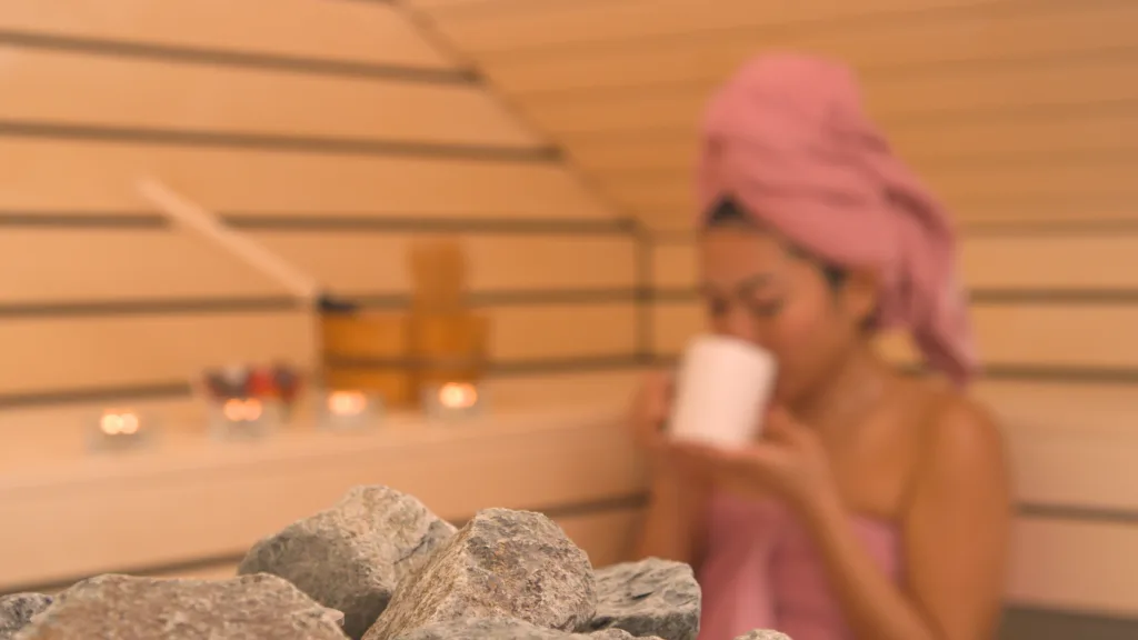 DOF = DEPTH OF FIELD: Sauna stones and young woman enjoying tea in background. Living healthy lifestyle for overall wellbeing. Beautiful Asian lady drinking hot cup of tea and relaxing in spa centre.