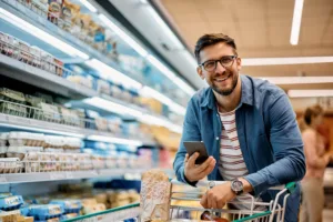 Happy man using smart phone while shopping in supermarket and looking at camera.