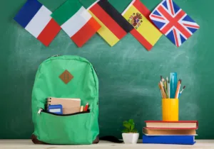 Learning languages concept - green backpack, flags of Spain, France, Great Britain and other countries, books and school supplies on the background of the blackboard