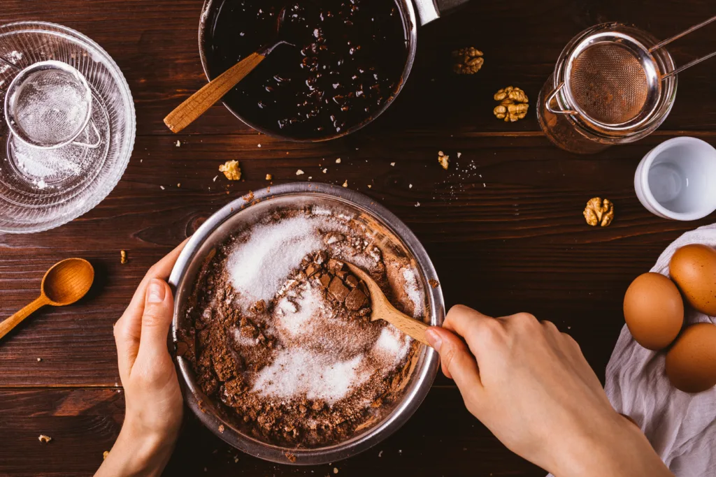 Top view female hands mix cocoa powder, sugar and flour to make dough with melted chocolate and walnuts for delicious homemade brownie cake.