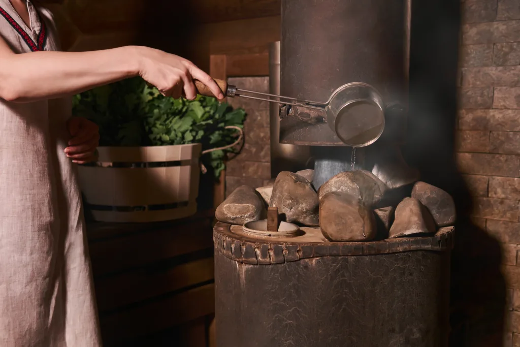 woman in traditional steam bath pours water on hot stones on stove