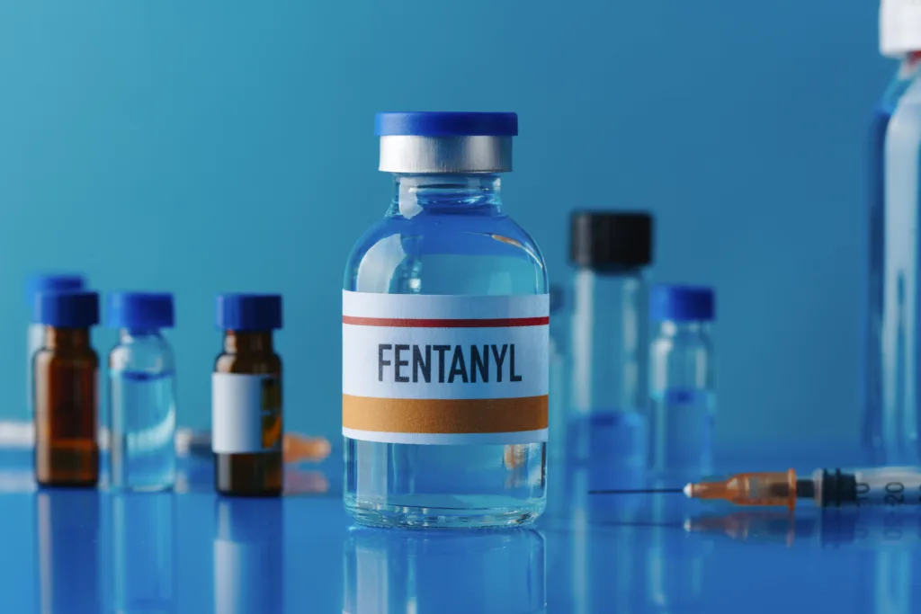 closeup of a simulated vial of fentanyl on a blue table next to a syringe and some other different vials