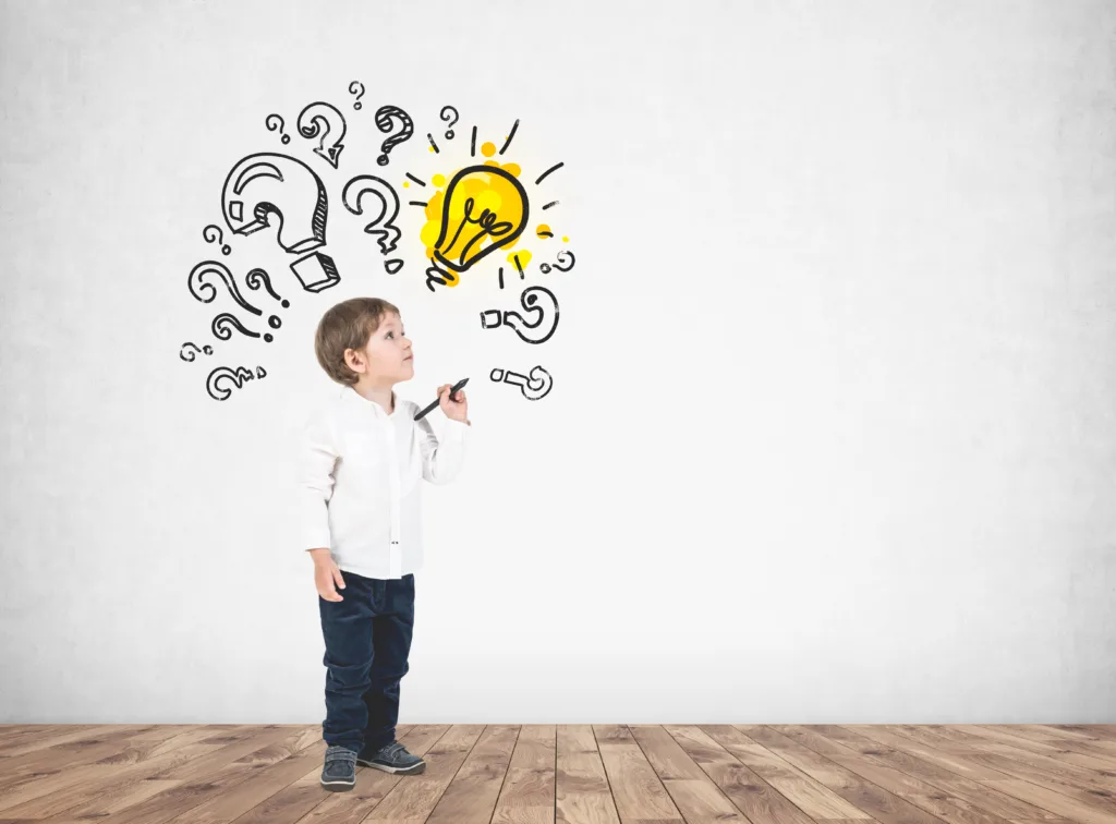 Cute little boy in a white shirt and dark jeans holding a marker and looking upwards. Question marks and a light bulb on a concrete wall. Mock up