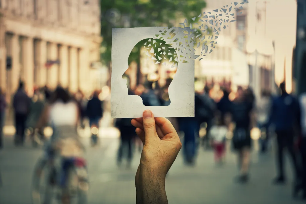 Hand holding a paper sheet with human head icon broken into pieces over a crowded street background. Concept of memory loss and dementia disease. Alzheimer's losing brain and memory function.