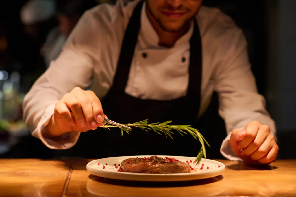 Hand of young male chef putting aromatic herb on top of roasted meat seasoned with spices while preparing tasty dish for serving