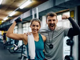 Happy athletic woman and her fitness instructor flexing muscles in a gym while looking at camera.