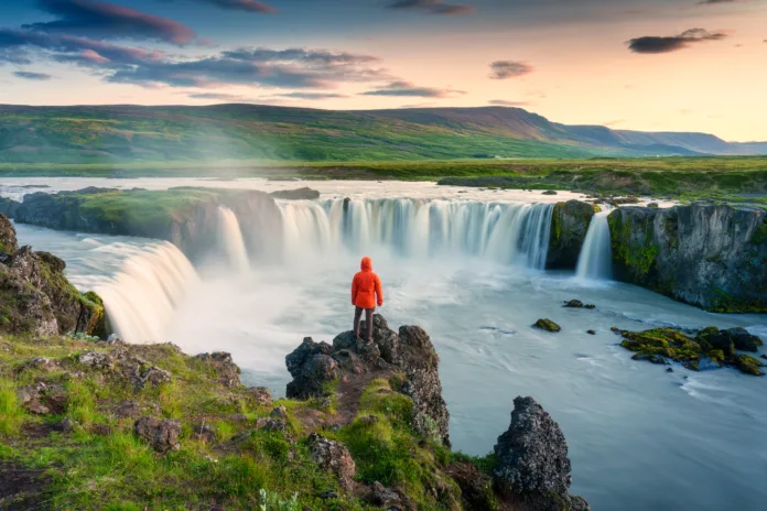 Majestic landscape of Godafoss waterfall flowing with colorful sunset sky and male tourist standing at the cliff on Skjalfandafljot river in summer at Northern Iceland