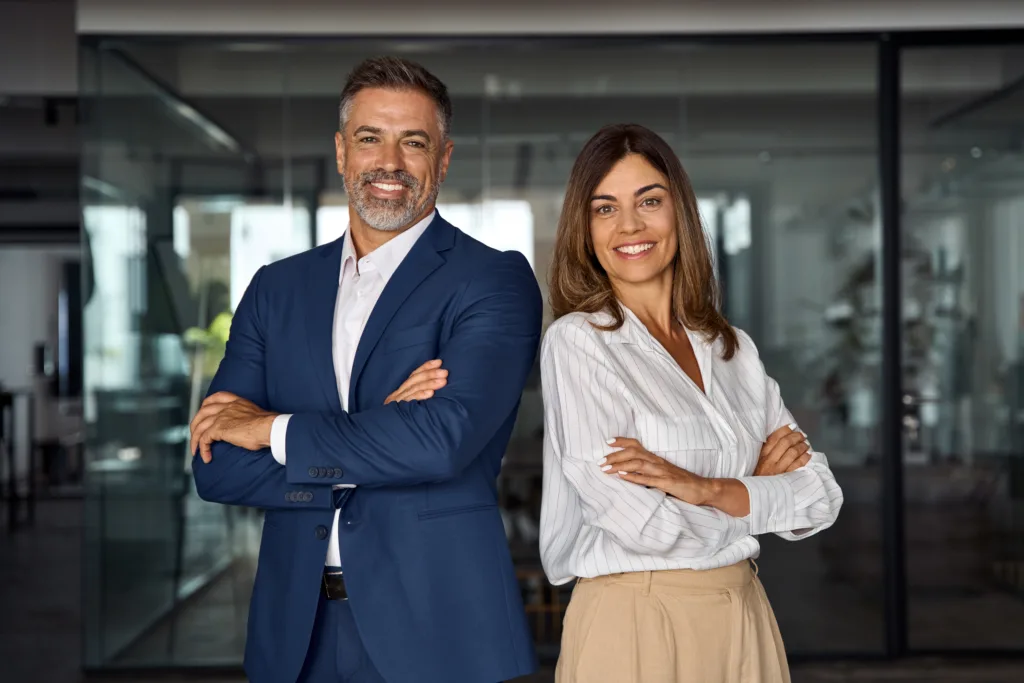 Portrait of smiling mature Latin or Indian business man and European business woman standing arms crossed in office. Two diverse colleagues, group team of confident professional business people.