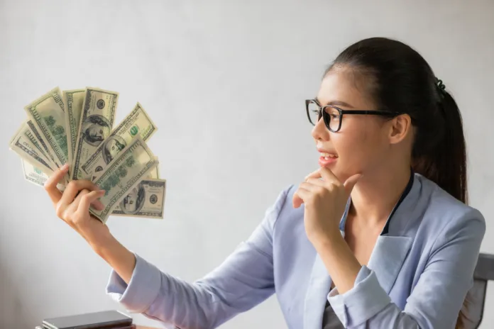 Successful Asian business young woman holding money US dollar bills in hand on office desk, business finance concept