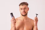 Young confused shirtless man holding in one hand electric shaving machine and shaver in another while isolated on white background. Handsome man making hard decision. Male beauty and skin care concept