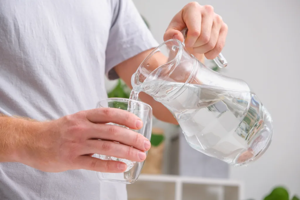 A man pours cold water into a glass. Close-up of male hands pouring water from a jug into a glass tumbler. A man quenches his thirst.