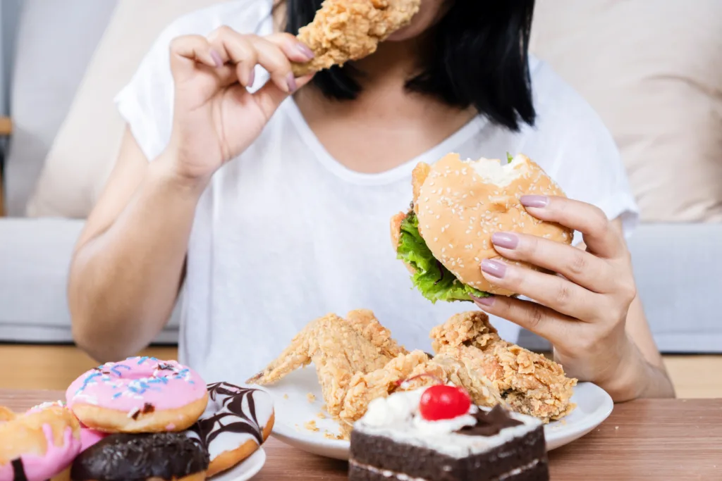 Binge eating disorder concept with woman eating fast food burger, fired chicken , donuts and desserts, over eating