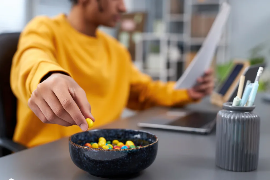 Close up of young man eating snacks while working or studying at home, focus on hand holding colorful candy, copy space