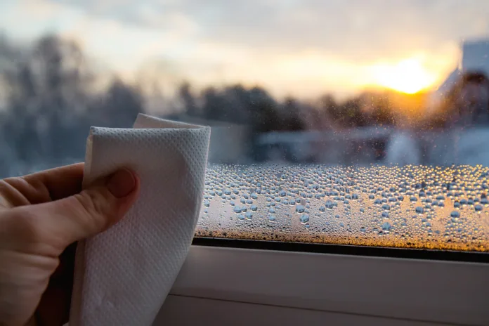 Close up view of person hand using paper cloth, drying wet condensation drops from glass window in cold winter morning at sunrise.