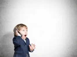 Cute little child is talking on his smartphone while standing in his business suit near a concrete wall. Mock up