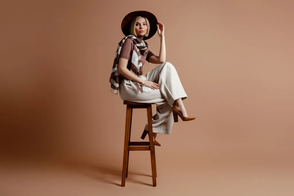 Fashionable young woman adjusting her elegant hat while sitting on a chair against brown background