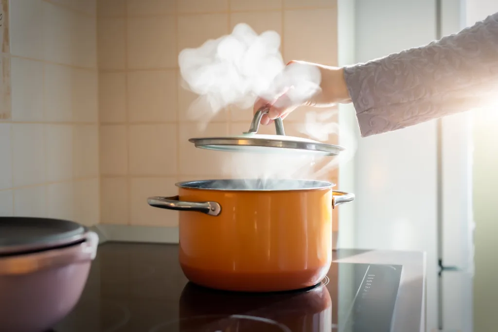 Female hand open lid of enamel steel cooking pan on electric hob with boiling water or soup and scenic vapor steam backlit by warm sunlight at kitchen. Kitchenware utensil and tool at home background.