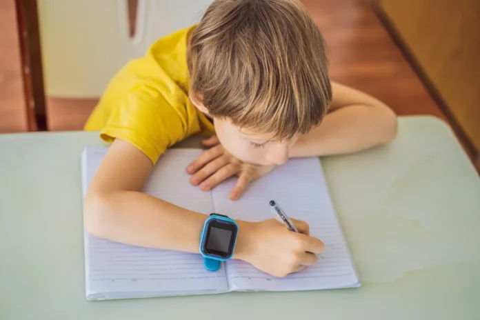 Little boy sitting at the table and looking smart watch. Smart watch for baby safety. The child makes school lessons, listening to music, calling friends.