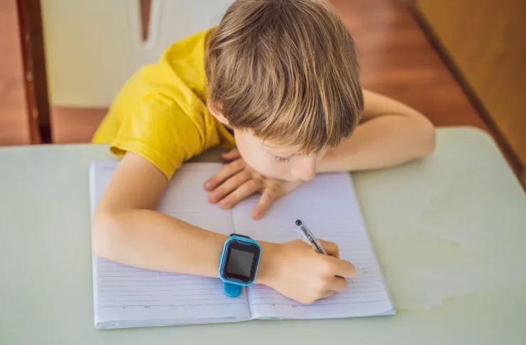 Little boy sitting at the table and looking smart watch. Smart watch for baby safety. The child makes school lessons, listening to music, calling friends.