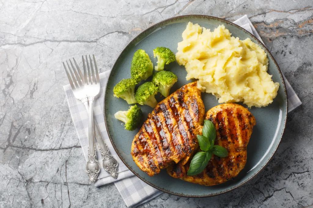 portion of grilled chicken breast with a side dish of mashed potatoes and broccoli close-up in a plate on the table. Horizontal top view from above