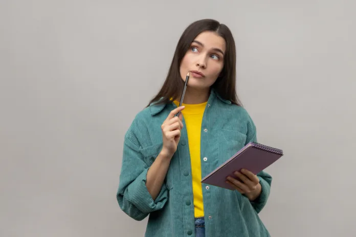 Thoughtful woman standing with paper notebook and pen, looking away with pensive expression, making plan, wearing casual style jacket. Indoor studio shot isolated on gray background.