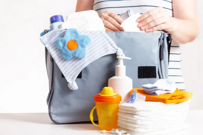 Unrecognizible pregnant caucasian woman in striped t-shirt packing big blue diaper bag to maternity hospital. Diapers, nappy, hat, bottle and other necessary things for newborn baby.