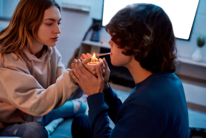 Young girl lighting cannabis cigarette for her boyfriend while sitting on the couch at home. Young couple smoking marijuana joint and relaxing. Marijuana, drugs, cannabis and weed legalization