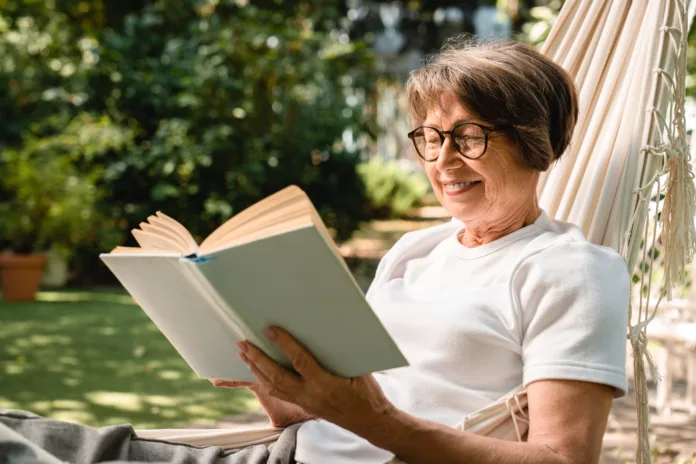 Happy relaxed old senior elderly woman grandmother relaxing while reading book in hammock outdoors in garden forest. Spending time outside