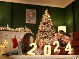 Parents and children having fun celebrating New Year together at home, lying by the Christmas tree holding illuminative numbers 2024