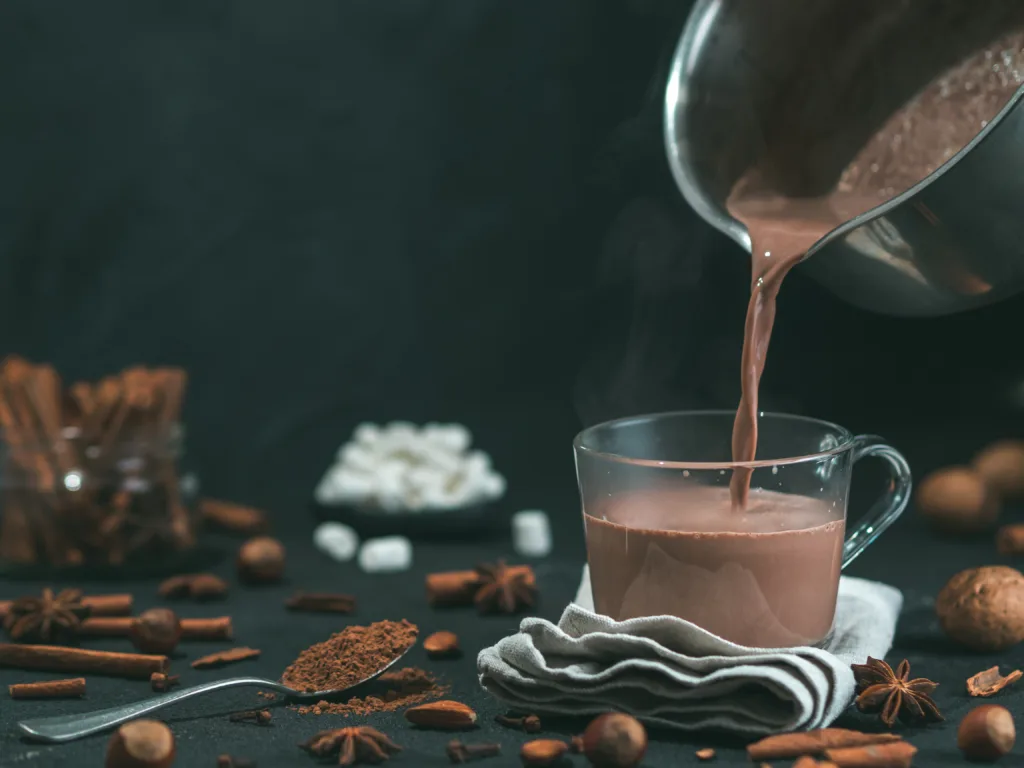 Pouring tasty hot chocolate cocoa drink into glass mug with ingredients on black table. Copy space Dark background. Low key.