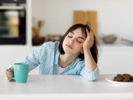 Sleepy young woman drinking coffee, feeling tired, suffering from insomnia and sleeping disorder. Sad female sitting in modern kitchen interior, empty space