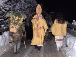 This image depicts a group of Krampus figures walking through a snowy village in Austria. The figures are dressed in traditional Krampus costumes, with horns, fur, and bells.